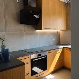 Honesto - Jasna kuchnia z drewnianymi frontami szafek, czarnym blatem i sprzętem AGD w odcieniach szarości, oświetlona promieniami słońca padającymi na podłogę.