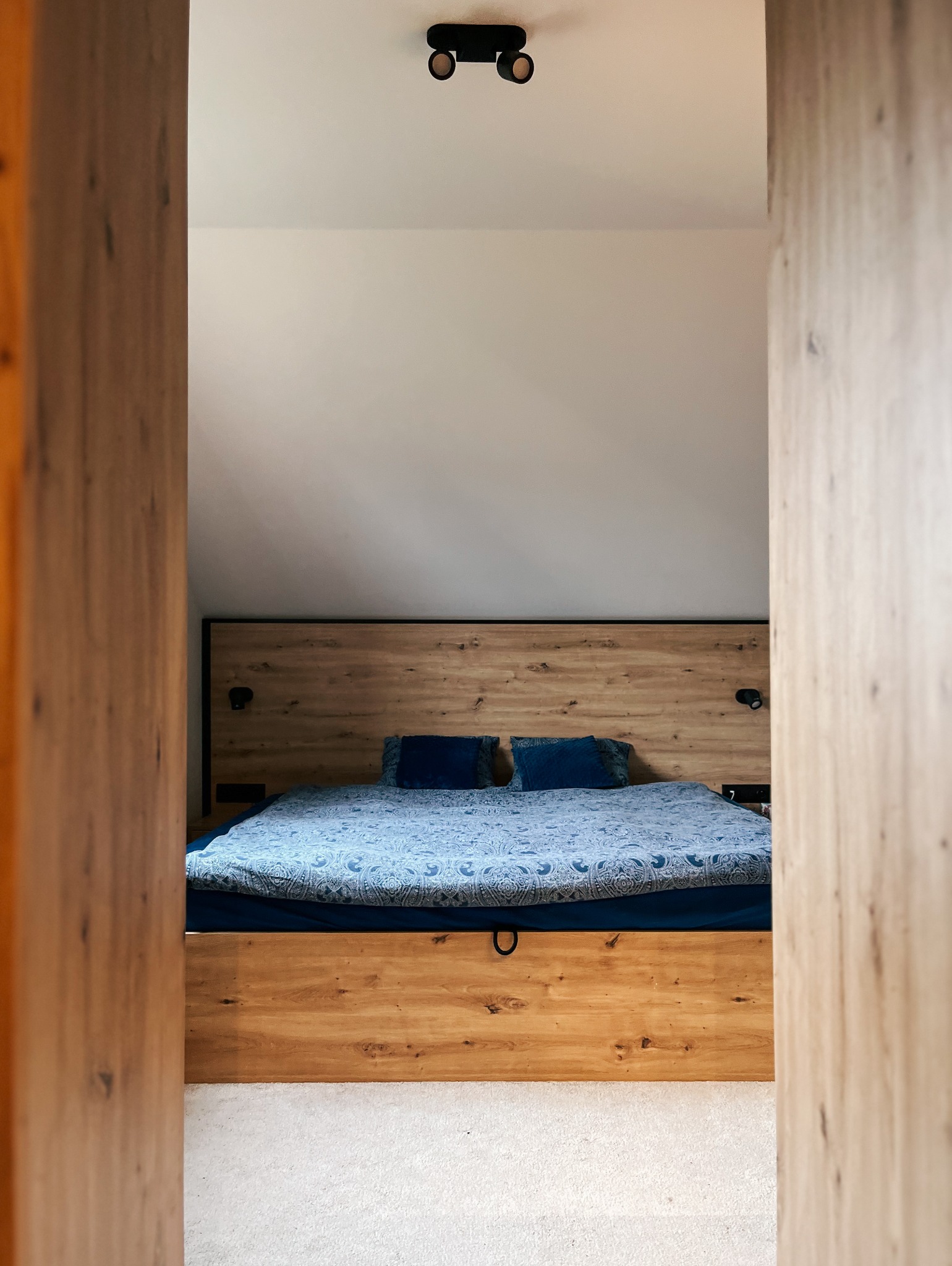 Drewniane łóżko z wezgłowiem wkomponowane w skos dachu, widoczne przez drewniane drzwi, oświetlone punktowym światłem.