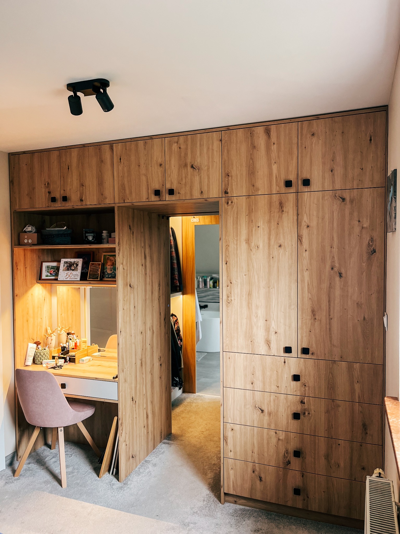 Zabudowa z drewna z szafami i biurkiem z oświetleniem, czarne kwadratowe uchwyty, widok na łazienkę w tle.