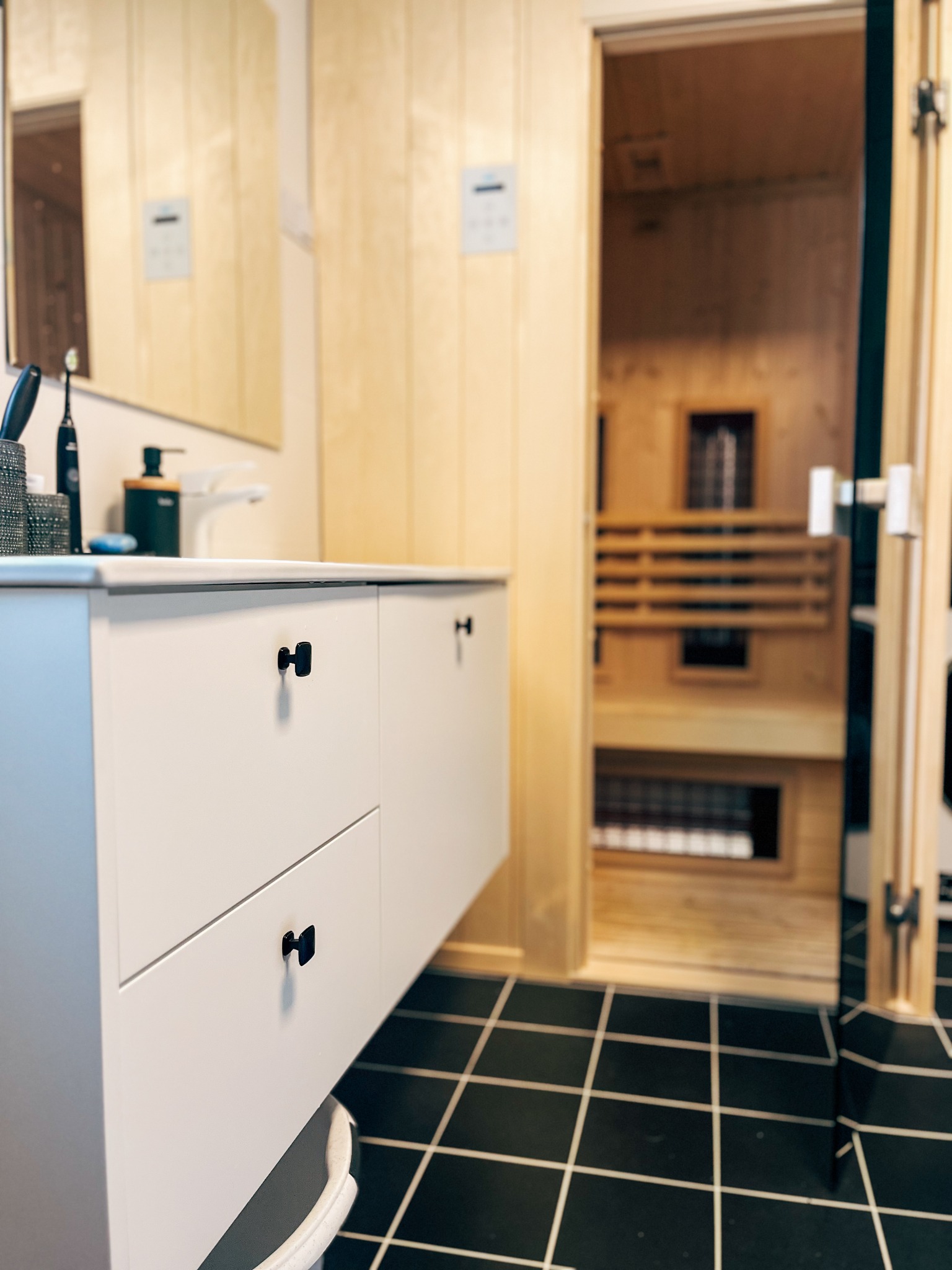 Nowoczesna łazienka z białą szafką pod umywalką z czarnymi uchwytami i widoczną sauną w tle.