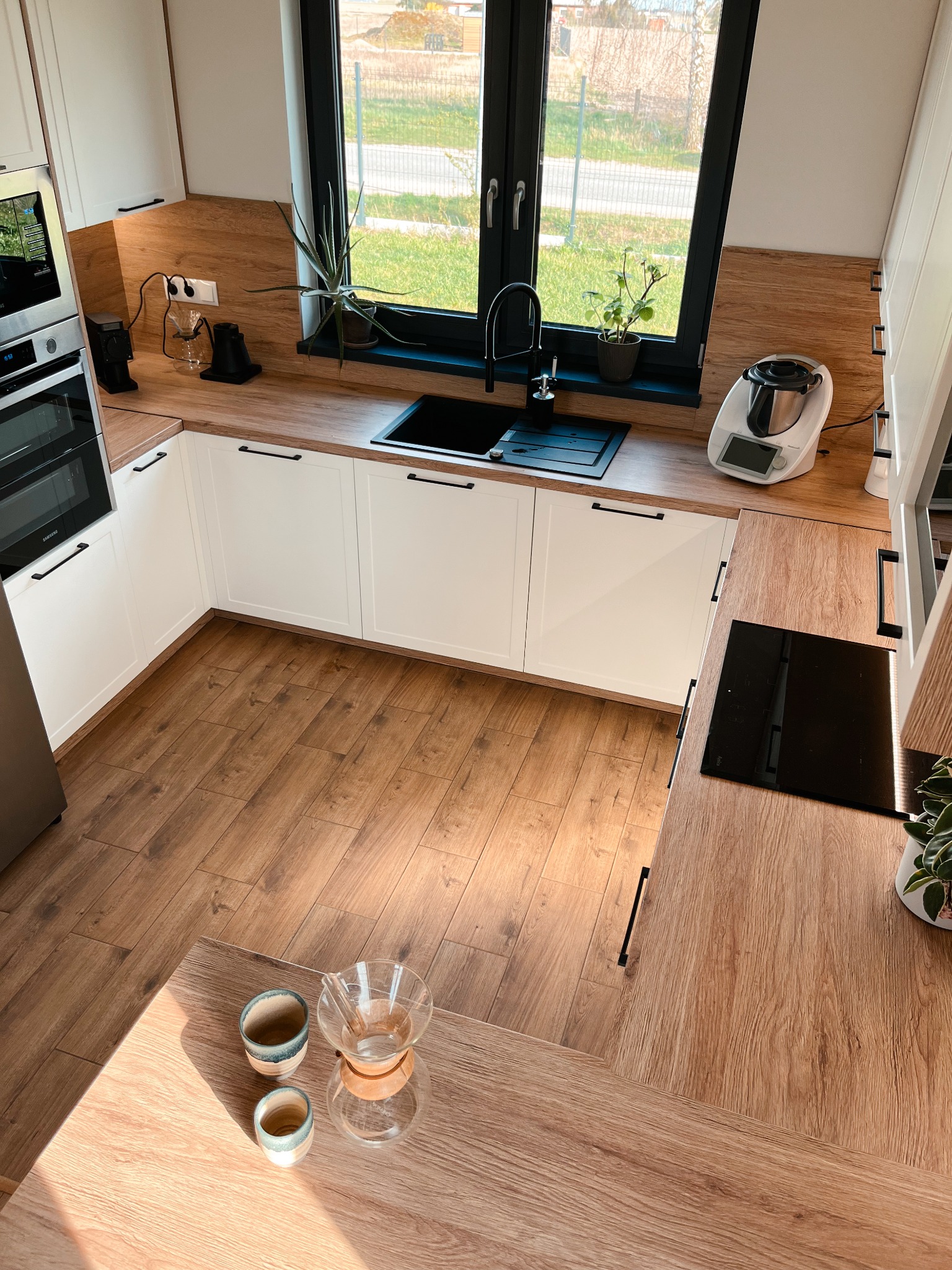 Nowoczesna kuchnia w jasnych barwach z drewnianymi akcentami, widok z góry, z czarnym oknem i wyposażeniem kuchennym.