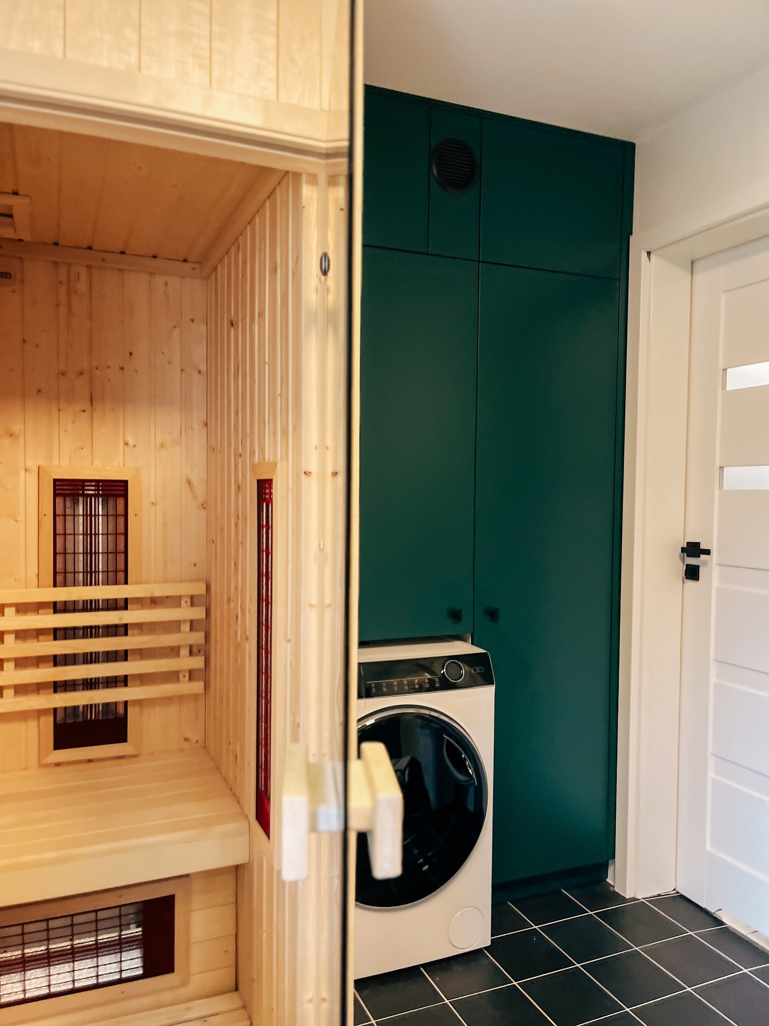 Wnętrze z sauną z jasnego drewna, obok ciemnozielona szafa w zabudowie, pralka i białe drzwi z czarną klamką na czarnej podłodze z płytek.