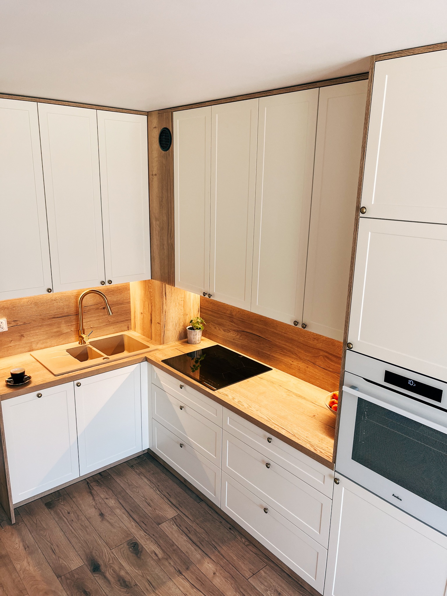 Nowoczesna kuchnia w odcieniach bieli i drewna z złotą armaturą, czarną płytą indukcyjną i drewnianą podłogą, widziana z góry.