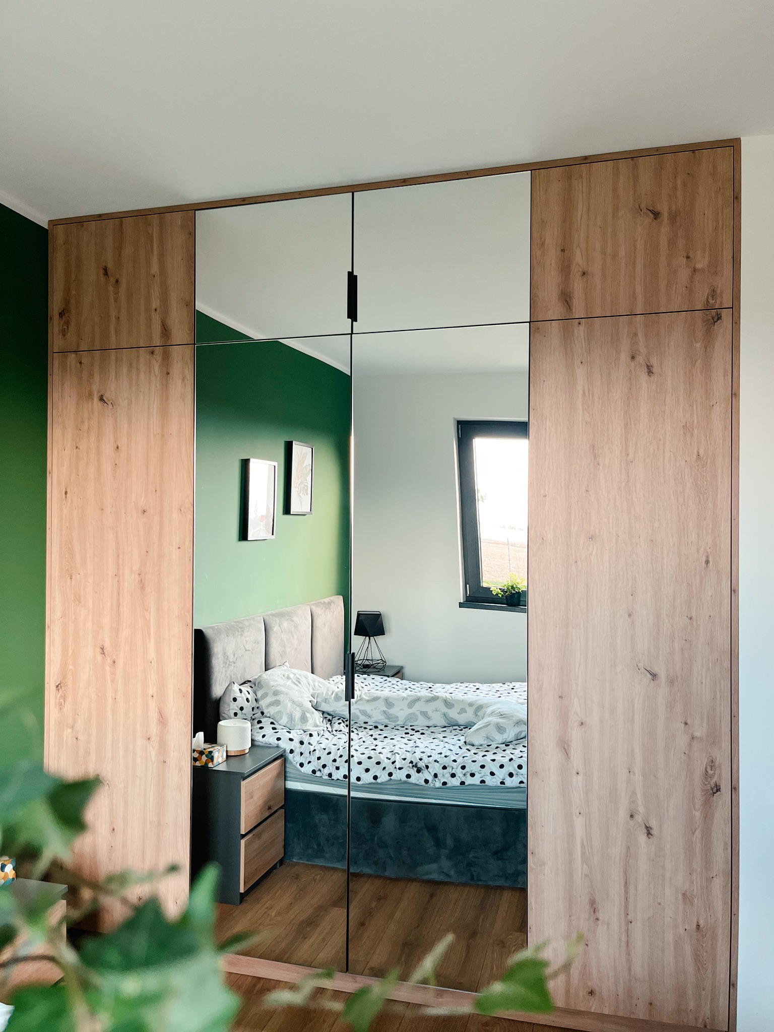 Szafa wnękowa z lustrzanymi drzwiami przesuwnymi i drewnianymi bokami, odbijająca sypialnię z zieloną ścianą i łóżkiem w kropki.