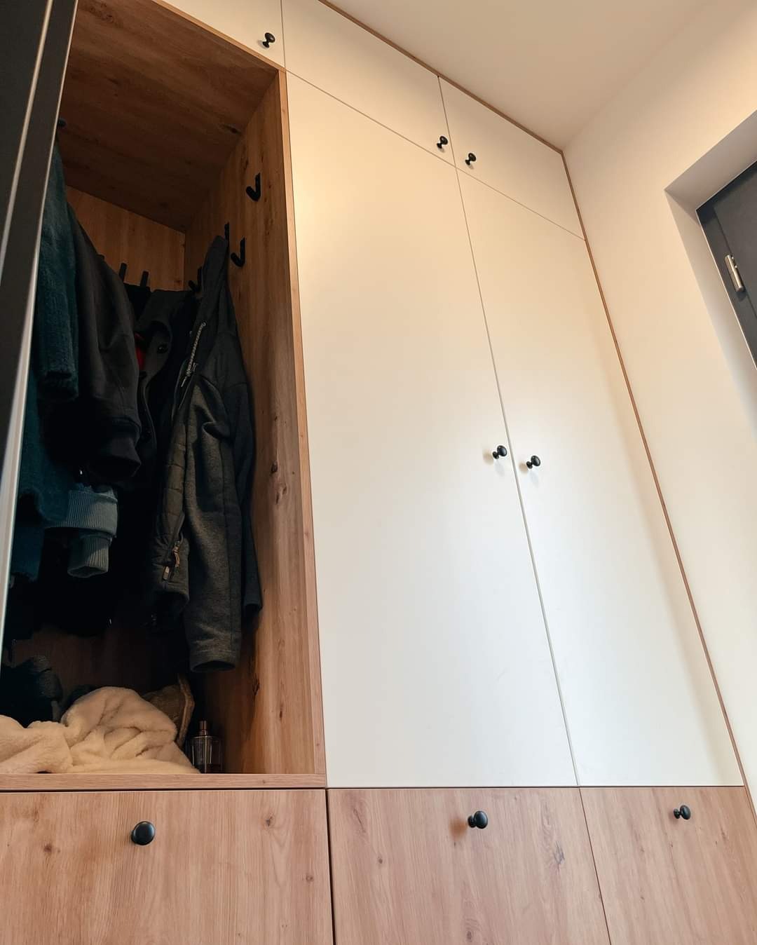 Wysoka szafa wnękowa z białymi frontami i drewnopodobnymi dolnymi szafkami, z otwartą częścią z wieszakami na ubrania i perfumami na półce.