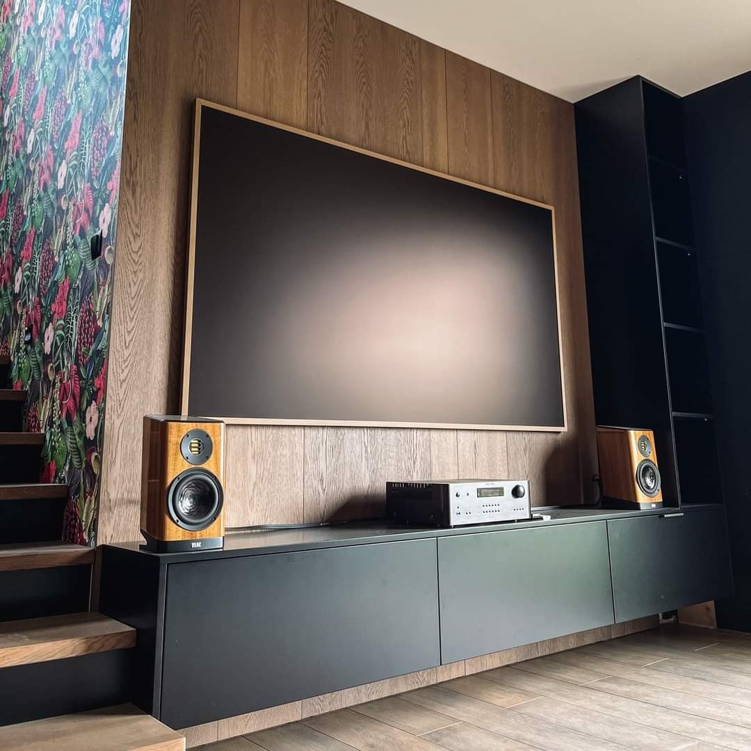 Nowoczesny salon z drewnianymi panelami ściennymi, czarną szafką RTV, głośnikami i dużym ekranem, schody z kwiatową tapetą po lewej stronie.