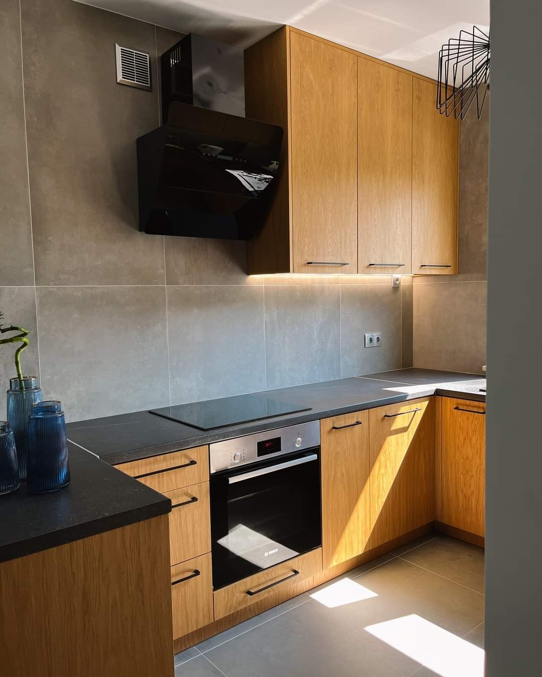 Jasna kuchnia z drewnianymi frontami szafek, czarnym blatem i sprzętem AGD w odcieniach szarości, oświetlona promieniami słońca padającymi na podłogę.