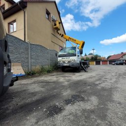 KlimAir Damian Szymański Montaż i Serwis klimatyzacji - Montaż klimatyzacji na elewacji domu z użyciem podnośnika koszowego, widoczny karton po klimatyzatorze marki Gree obok szarego ogrodzenia.