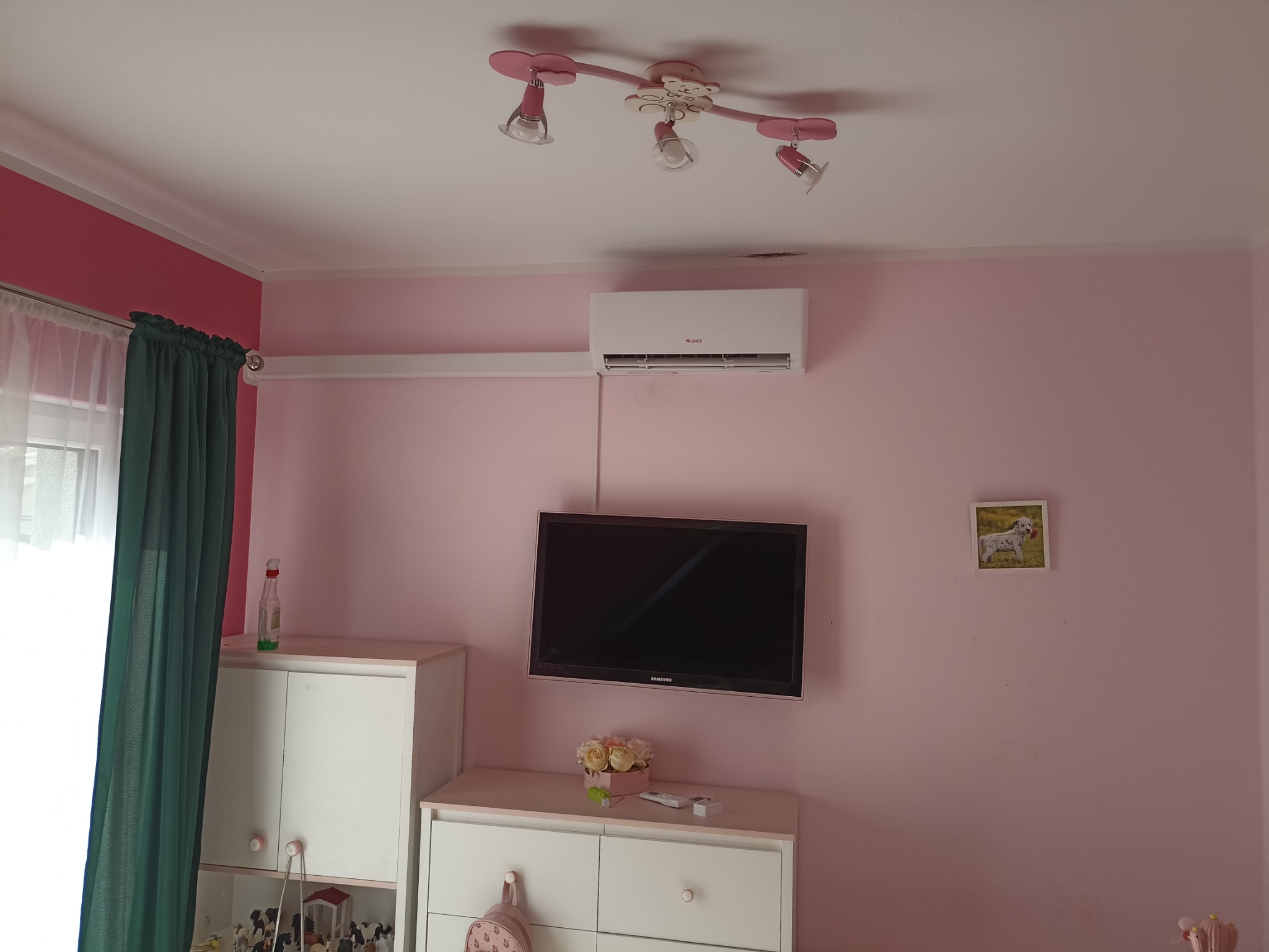 Klimatyzator marki Gree zamontowany na różowej ścianie w pokoju dziecięcym, widoczny telewizor, białe meble i lampa w kształcie kwiatków.