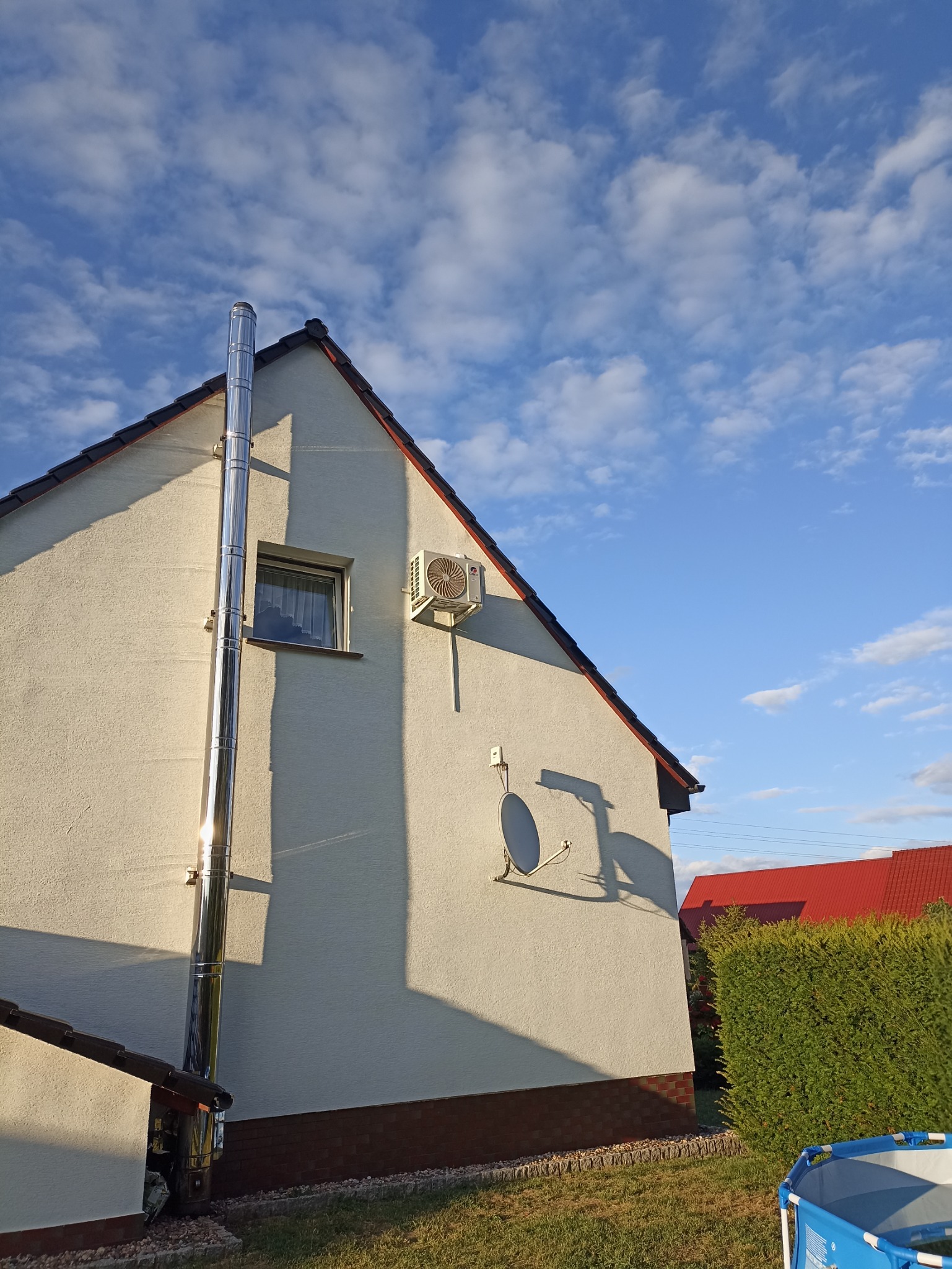 Biały dom jednorodzinny z zamontowaną klimatyzacją na ścianie szczytowej oraz rurą odprowadzającą spaliny, widoczne również antena satelitarna i fragment niebieskiego basenu.