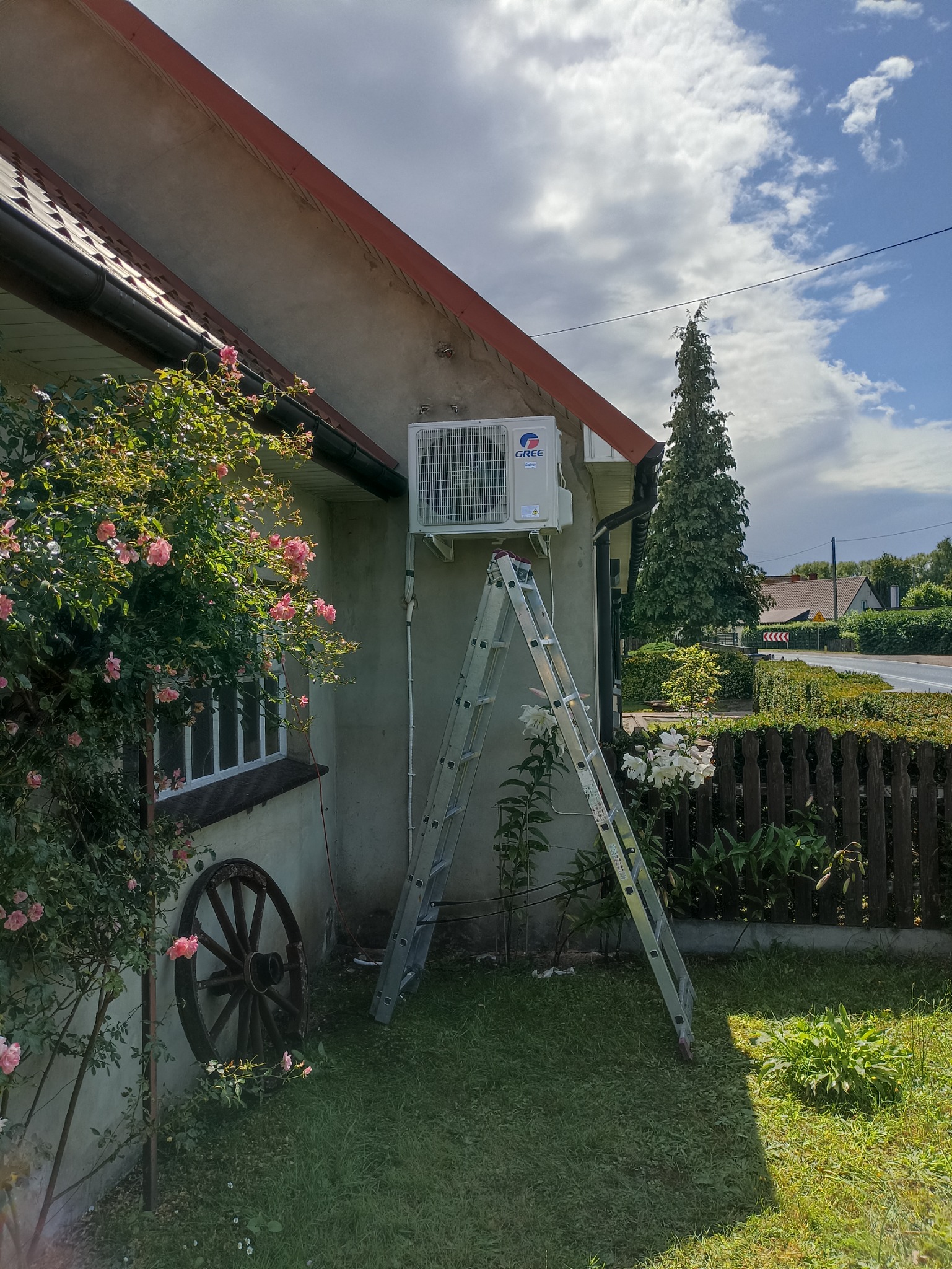 Biała jednostka zewnętrzna klimatyzatora marki Gree zamontowana na ścianie domu, z rozstawioną aluminiową drabiną, otoczona zielenią i kwiatami, w tle widoczny fragment ulicy.