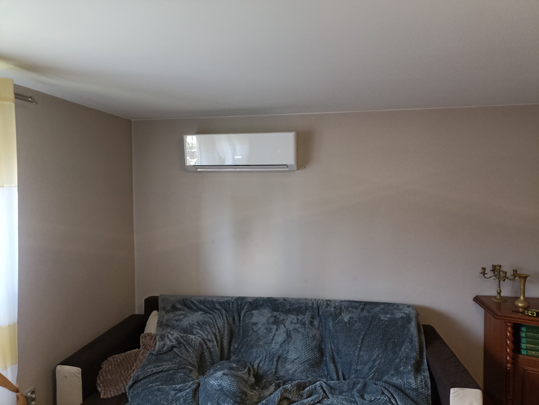 Nowoczesny klimatyzator zamontowany na ścianie w salonie we Wrocławiu, widoczny fragment sofy z narzutą i klasyczna komoda z dekoracyjnym świecznikiem.