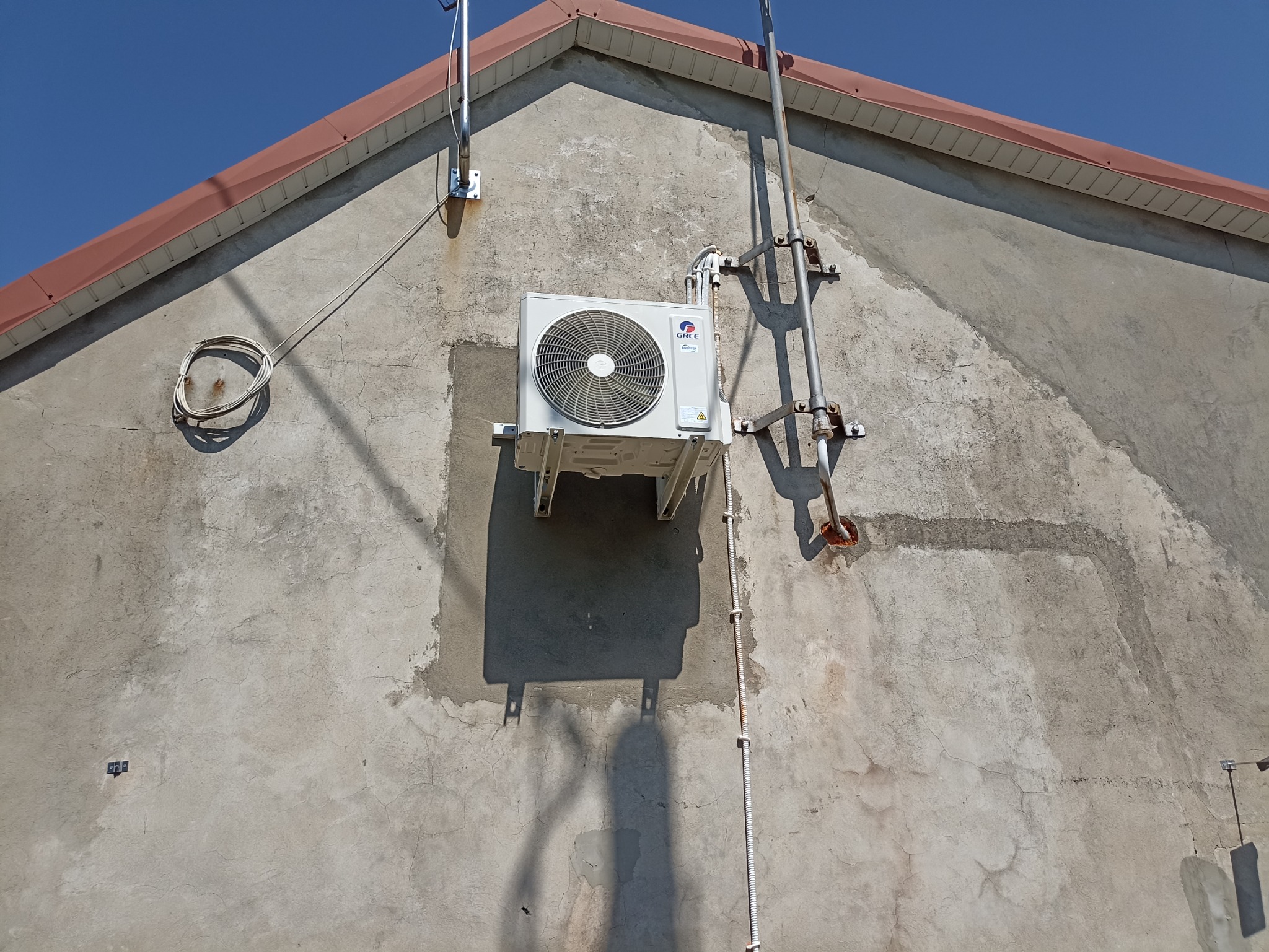 Zewnętrzna jednostka klimatyzacji marki Gree zamontowana na szarej, lekko popękanej ścianie budynku z widocznymi śladami po wcześniejszych pracach montażowych i instalacji.