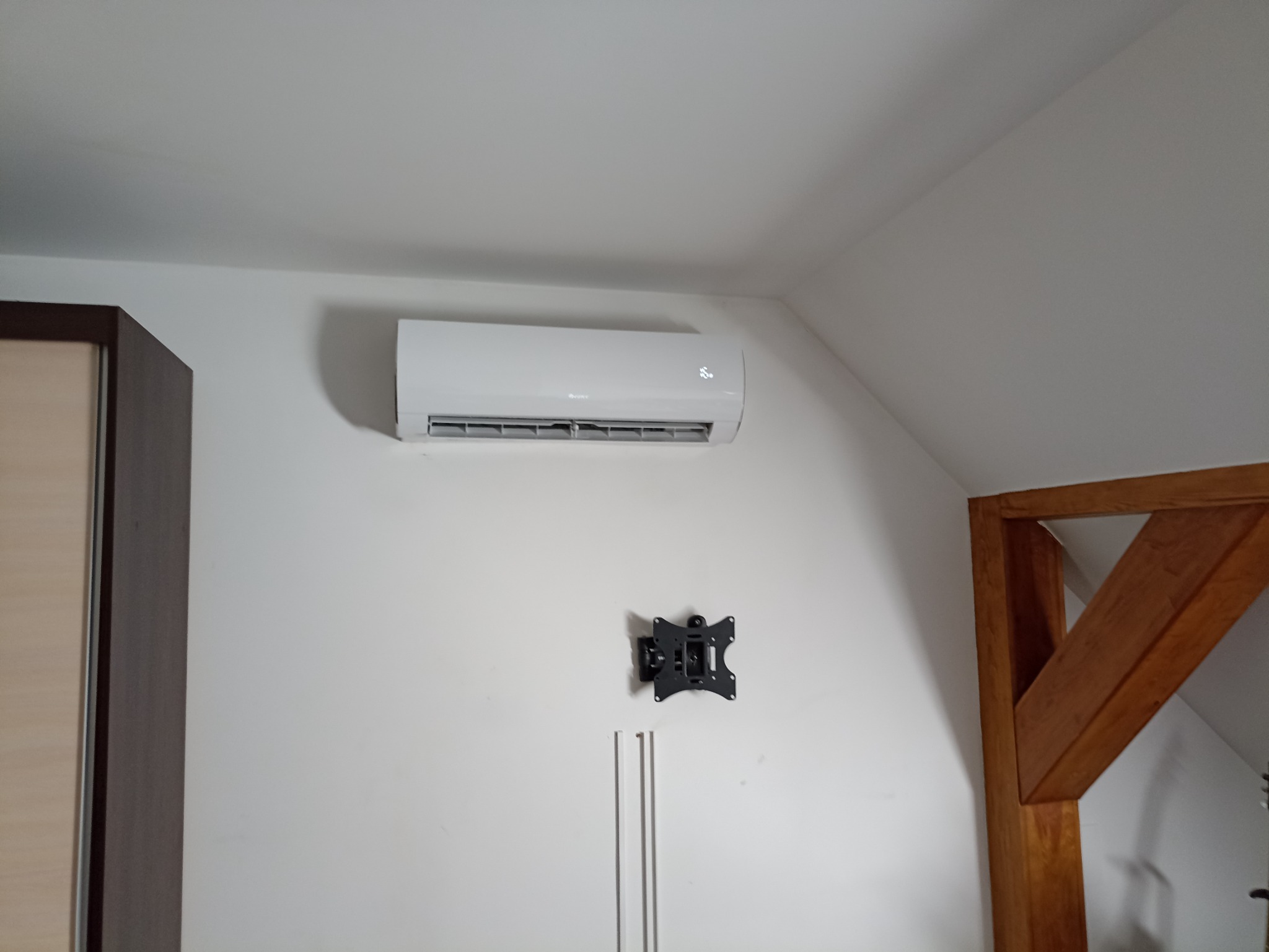 Zamontowany, biały klimatyzator na ścianie w pokoju na poddaszu. Widoczny również uchwyt do telewizora i drewniany element konstrukcyjny dachu.