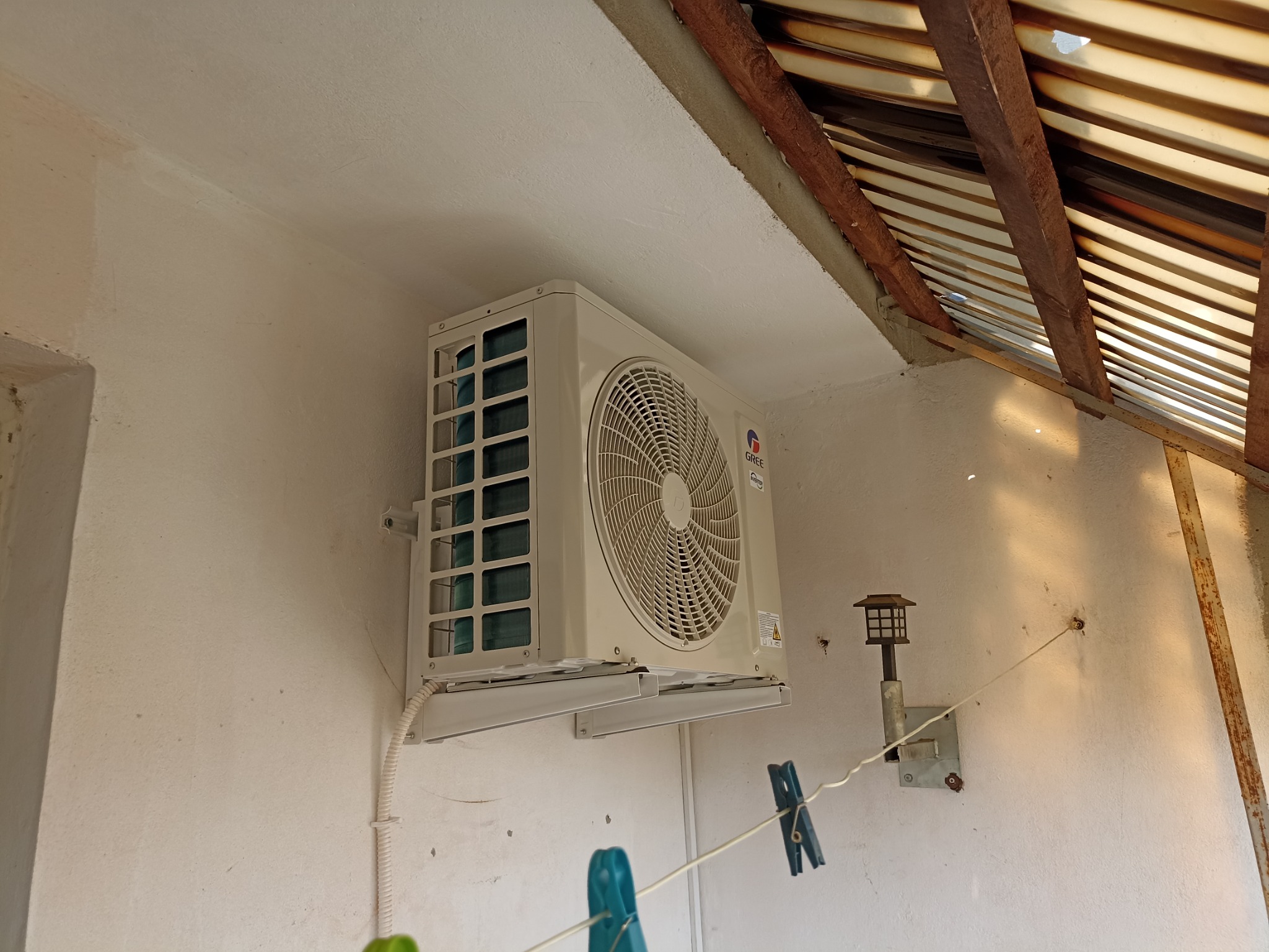 Zamontowany klimatyzator marki Gree na białej ścianie w pomieszczeniu z widocznym drewnianym dachem, obok lampa ścienna i sznurek z klamerkami.