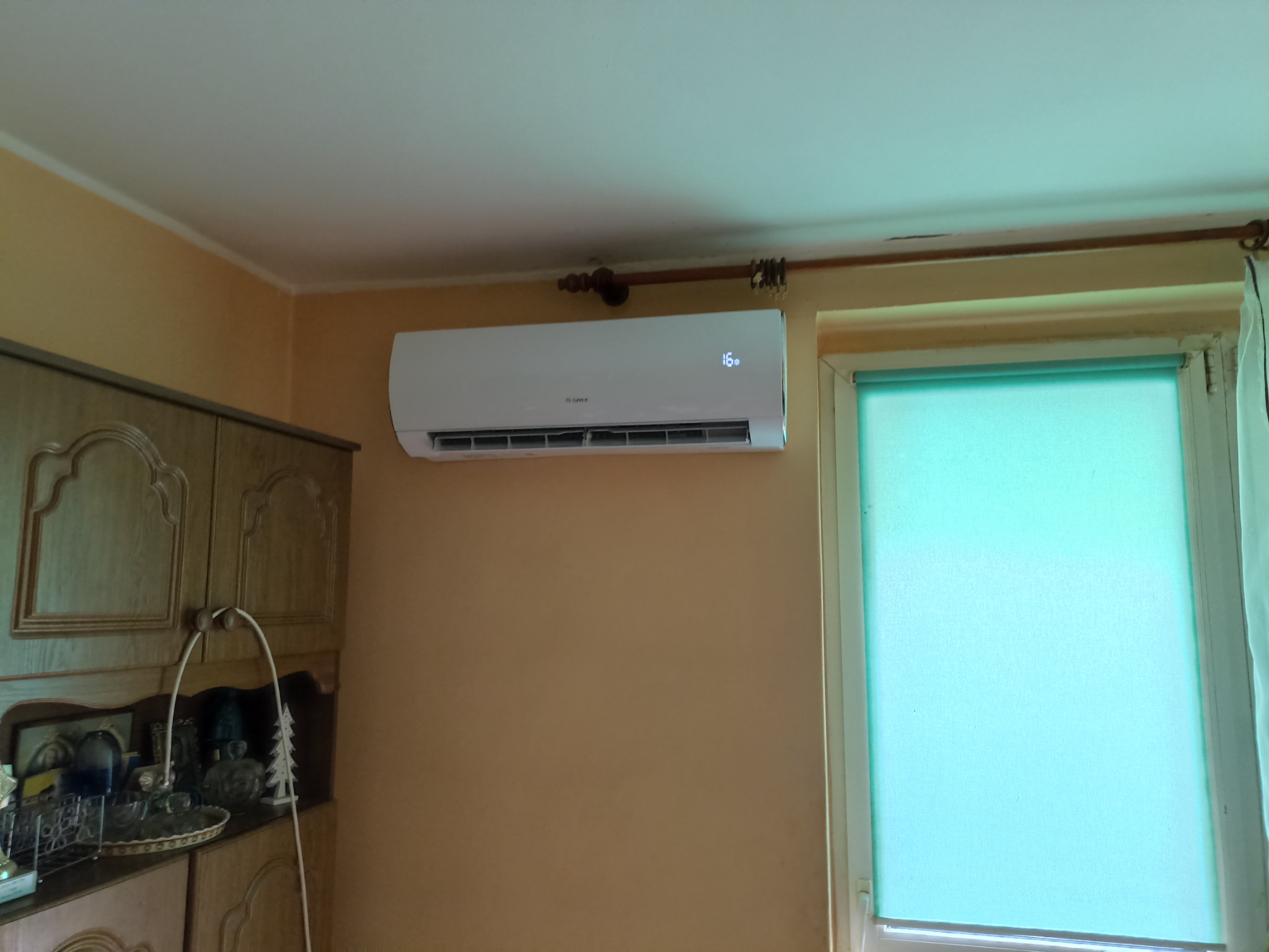 Biały klimatyzator marki Gree zamontowany na pomarańczowej ścianie w pokoju, obok okna z zieloną roletą i drewnianej szafki.