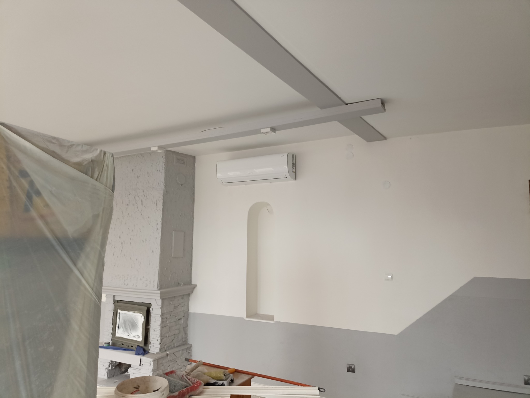 Biały klimatyzator zamontowany na ścianie w pomieszczeniu z kominkiem, widoczne szare belki dekoracyjne na suficie i elementy malarskie w dolnej części ściany, wnętrze w trakcie remontu, zakryte...