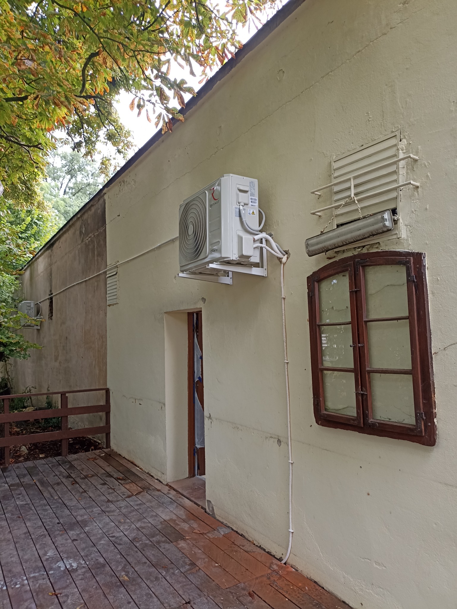 Zewnętrzna jednostka klimatyzacji marki Rotenso zamontowana na elewacji budynku z drewnianym oknem i podestem, widoczne przewody i rury instalacyjne.