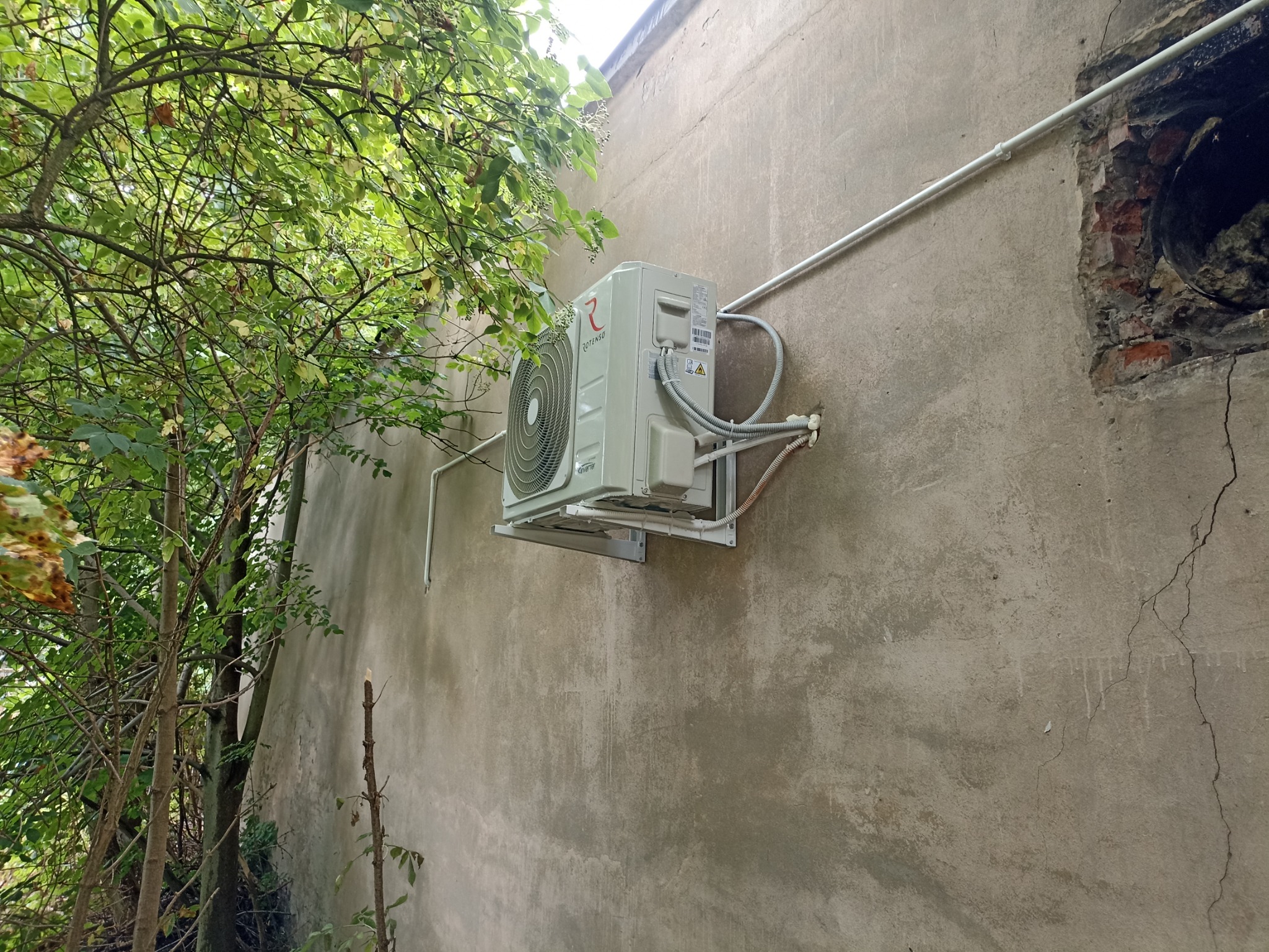 Zewnętrzna jednostka klimatyzacji Rotenso zamontowana na szarej, popękanej ścianie budynku, widoczna z boku, z fragmentem zieleni drzewa w kadrze.