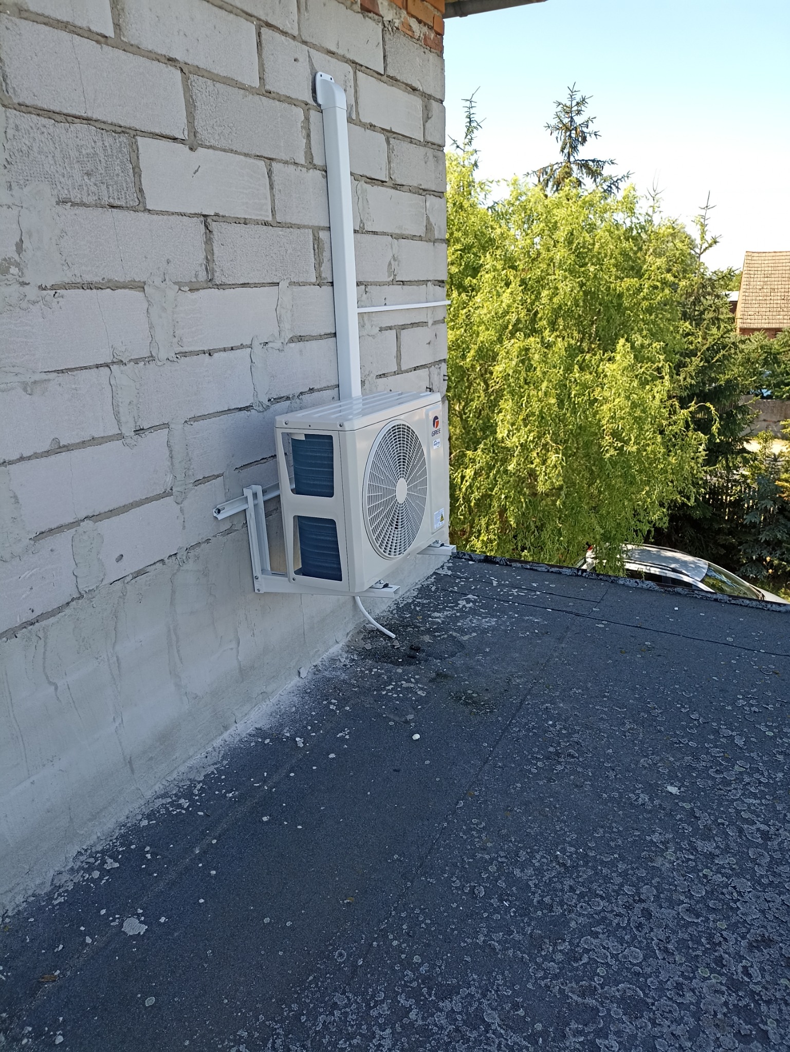 Zewnętrzna jednostka klimatyzacji marki Gree zamontowana na ścianie z białych bloczków, z widocznymi przewodami w białej maskownicy oraz fragmentem dachu pokrytego papą i zielenią w tle.