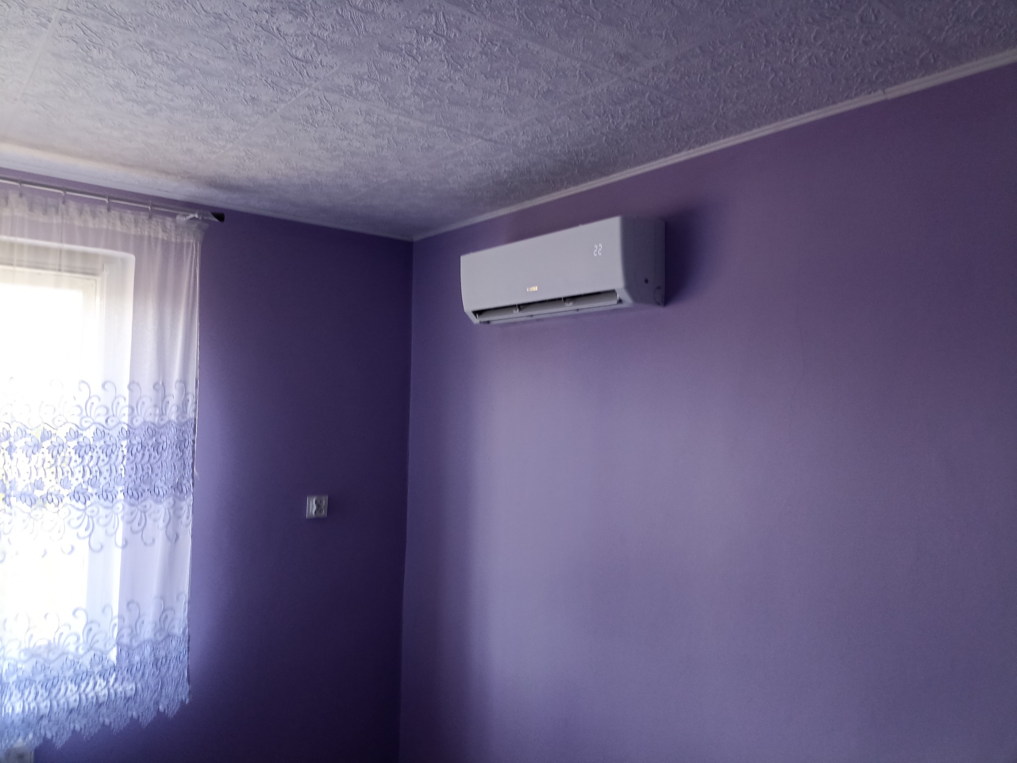 Biały klimatyzator ścienny zamontowany na fioletowej ścianie w pokoju z wzorzystym sufitem i oknem zasłoniętym firanką z haftem.