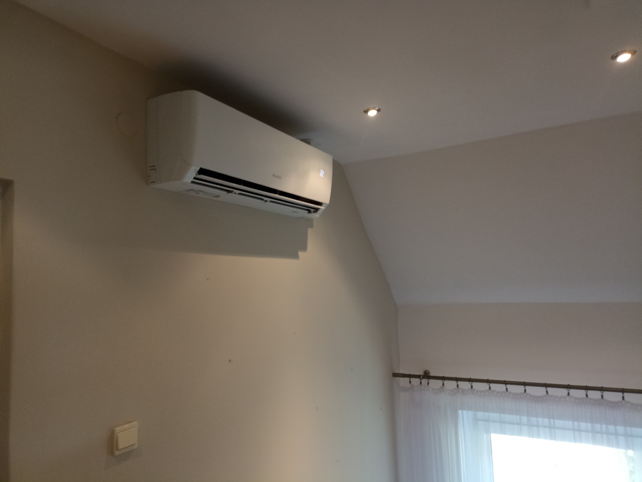 Zamontowany, biały klimatyzator ścienny z wyświetlaczem temperatury '22' w jasnym pomieszczeniu z beżowymi ścianami i punktowym oświetleniem sufitowym, widoczne firanki w oknie.