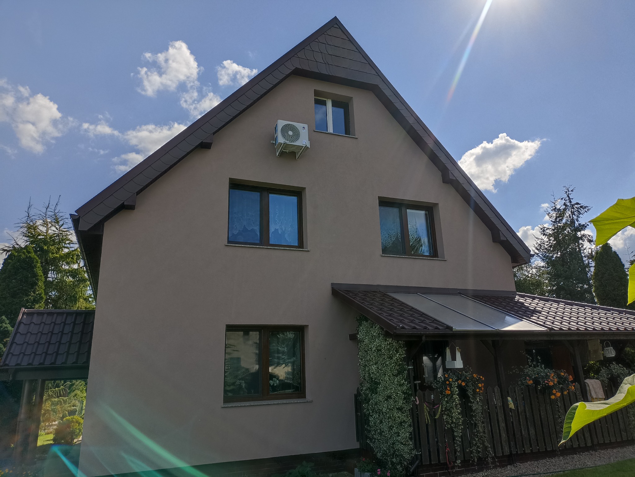 Dwupiętrowy dom z beżową elewacją i brązowym dachem, na ścianie zamontowany klimatyzator, widok z dołu w słoneczny dzień.