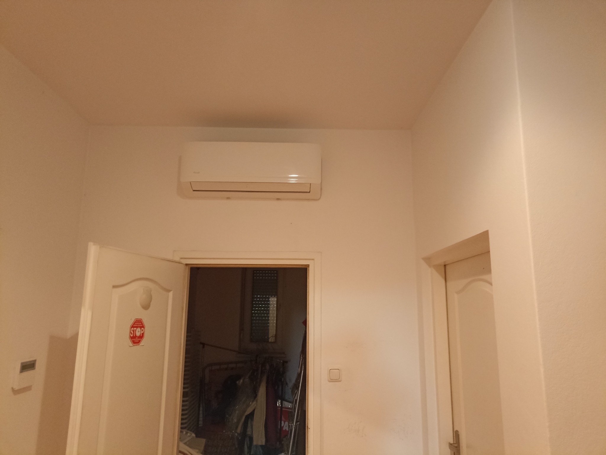 Biały klimatyzator ścienny zamontowany w pokoju z otwartymi drzwiami, na których widnieje naklejka 'STOP'.