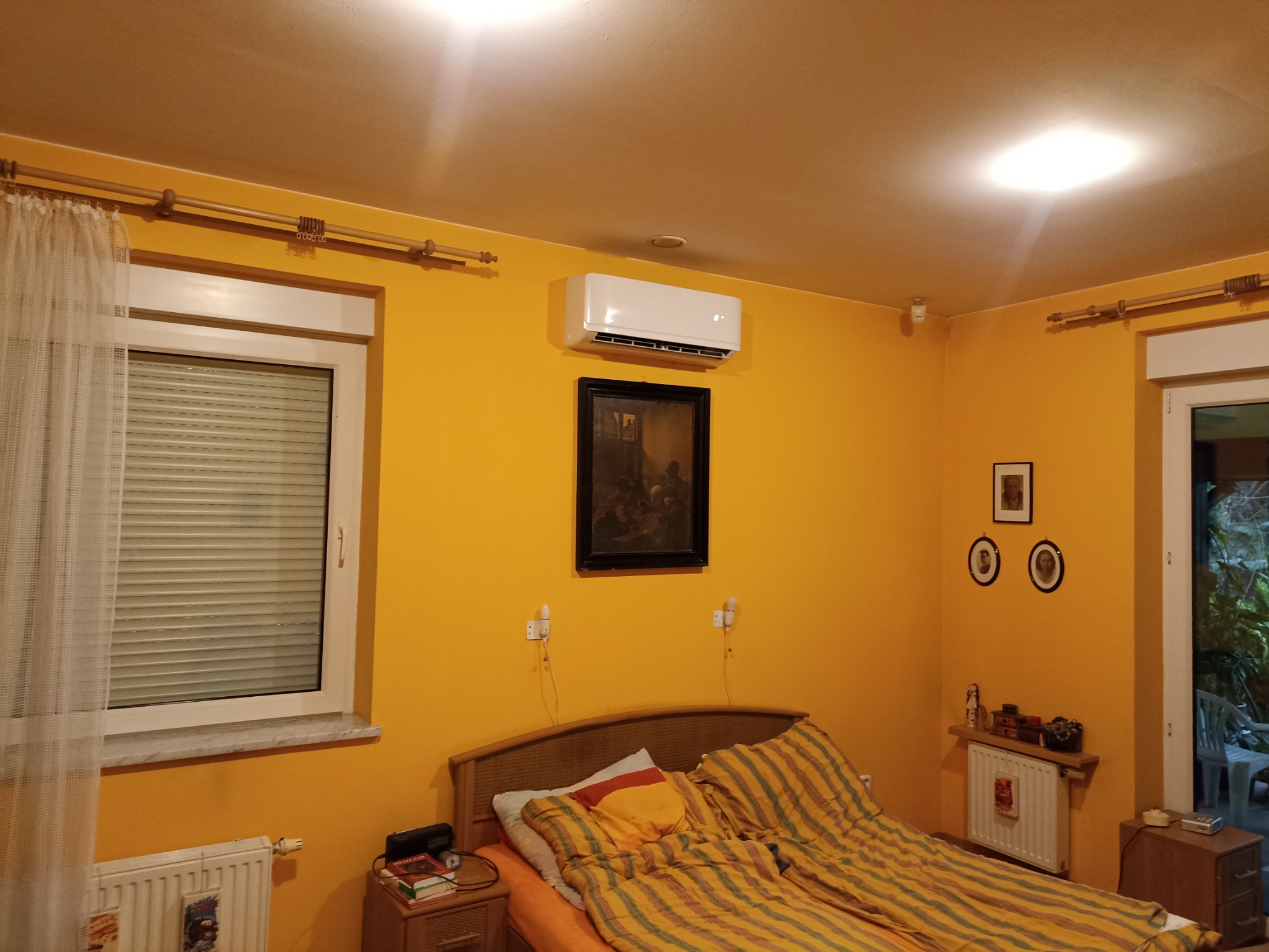 Żółty pokój z zainstalowaną białą klimatyzacją ścienną nad łóżkiem, pod nią wisi obraz w czarnej ramie, a po bokach lampki nocne.