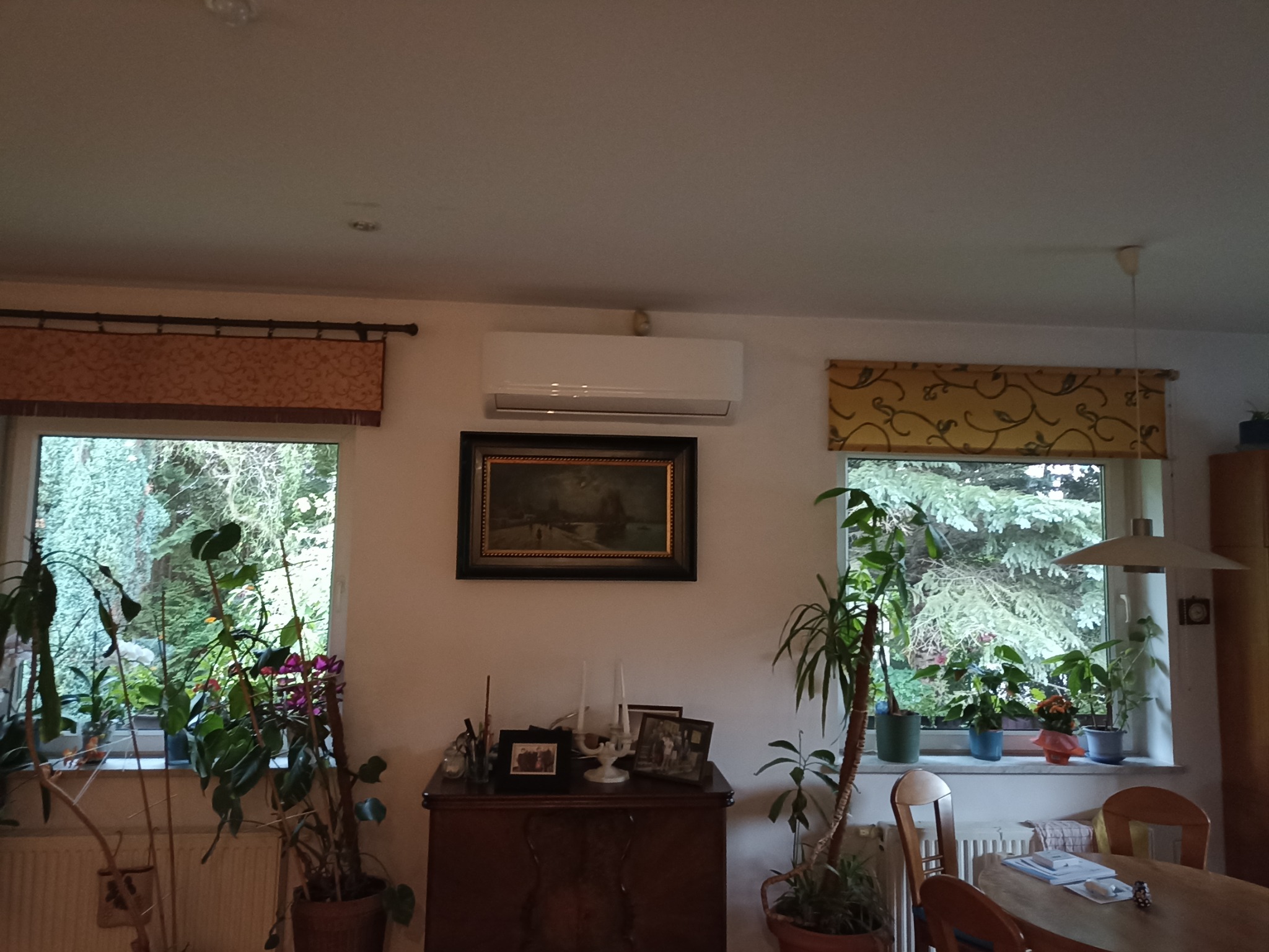 Biały klimatyzator ścienny zamontowany nad ciemną komodą z obrazem w ramie, widoczne dwa okna z roletami i roślinami doniczkowymi.