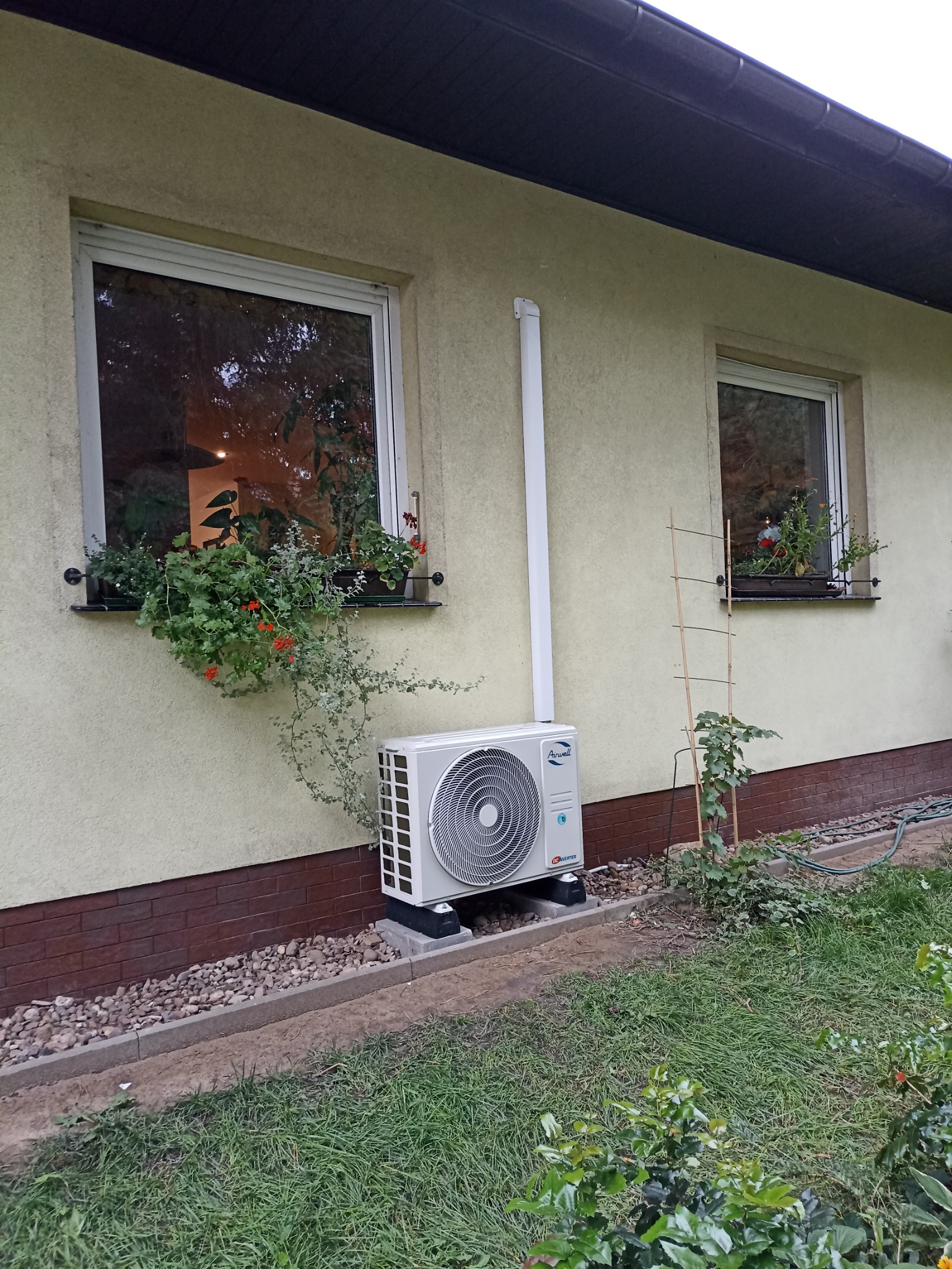 Zewnętrzna jednostka klimatyzacji zamontowana na elewacji domu, obok okna z kwitnącymi pelargoniami w skrzynkach. Widoczna rura odprowadzająca skropliny. Ściana pomalowana na jasnożółty kolor,...