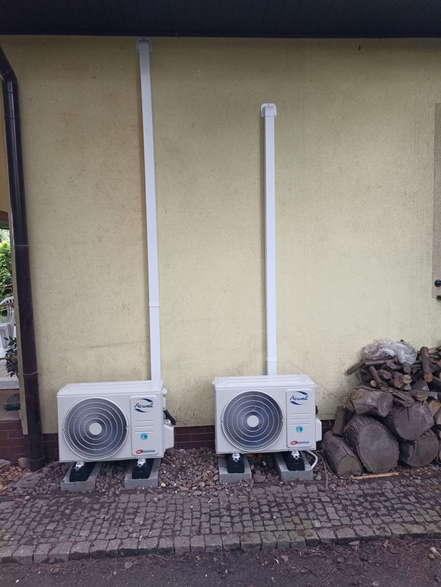 Dwa klimatyzatory Airwell zamontowane na zewnątrz budynku we Wrocławiu, z widocznymi rurami odprowadzającymi, ustawione na betonowych podstawach, obok stos drewna.