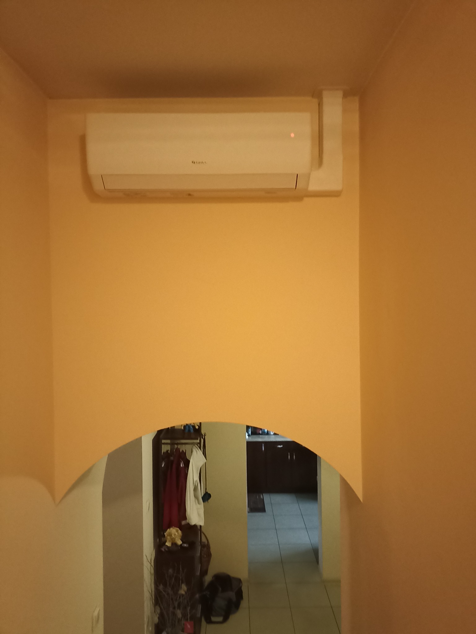 Biały klimatyzator ścienny zamontowany na pomarańczowej ścianie, widoczny przez łukowate przejście do innego pomieszczenia.