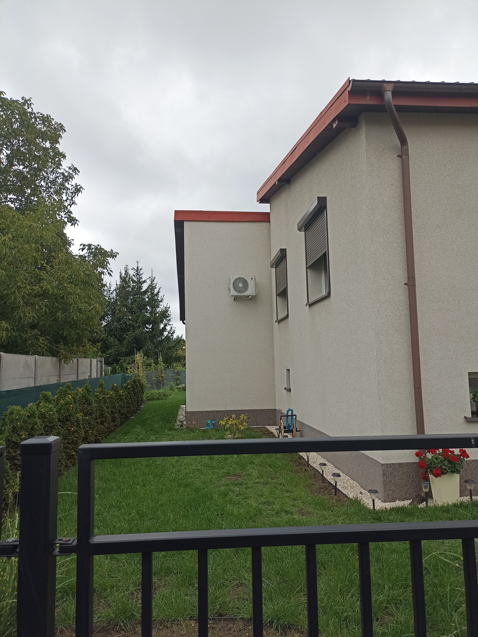 Zewnętrzna jednostka klimatyzacji zamontowana na ścianie domu z brązowymi roletami okiennymi, widoczna zza czarnego metalowego ogrodzenia w pochmurny dzień.