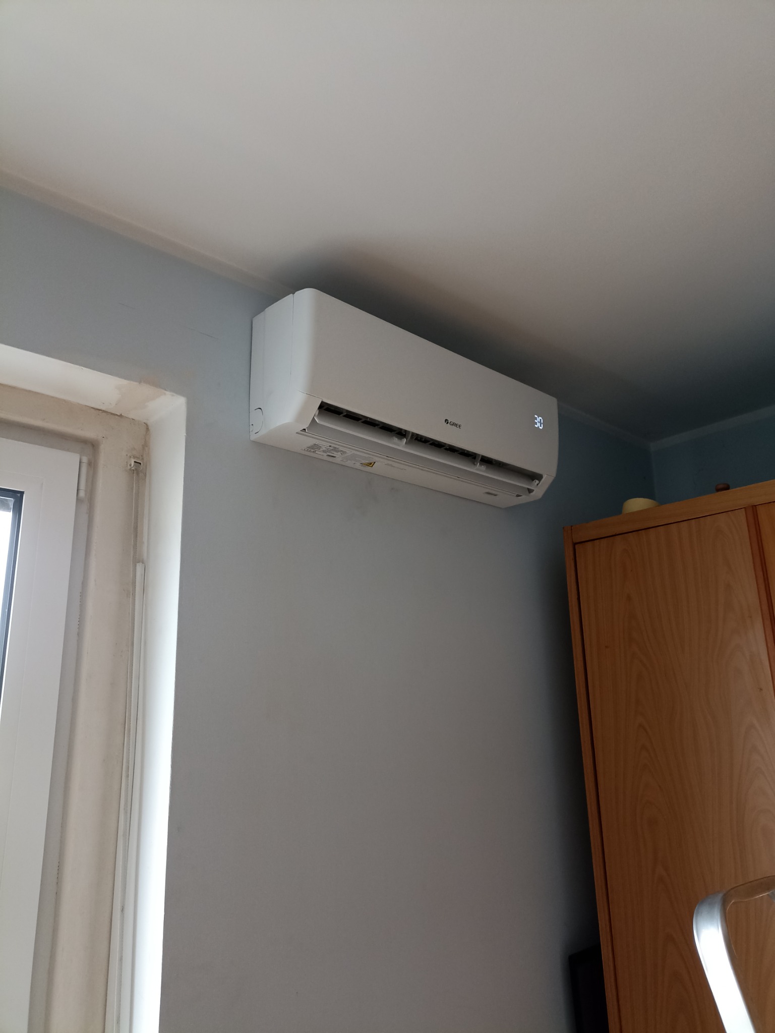 Biały klimatyzator marki Gree zamontowany na szarej ścianie w pomieszczeniu, widoczny wyświetlacz temperatury ustawiony na 30 stopni, obok okno z niedokończonym wykończeniem i drewniana szafa.