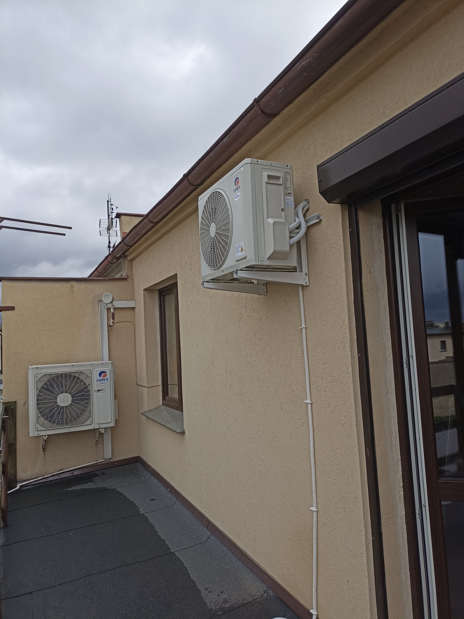 Dwa klimatyzatory marki Gree zamontowane na elewacji budynku z beżowym tynkiem strukturalnym, widoczne przewody w białej listwie, okno z brązową ramą i brązowy system rynnowy.