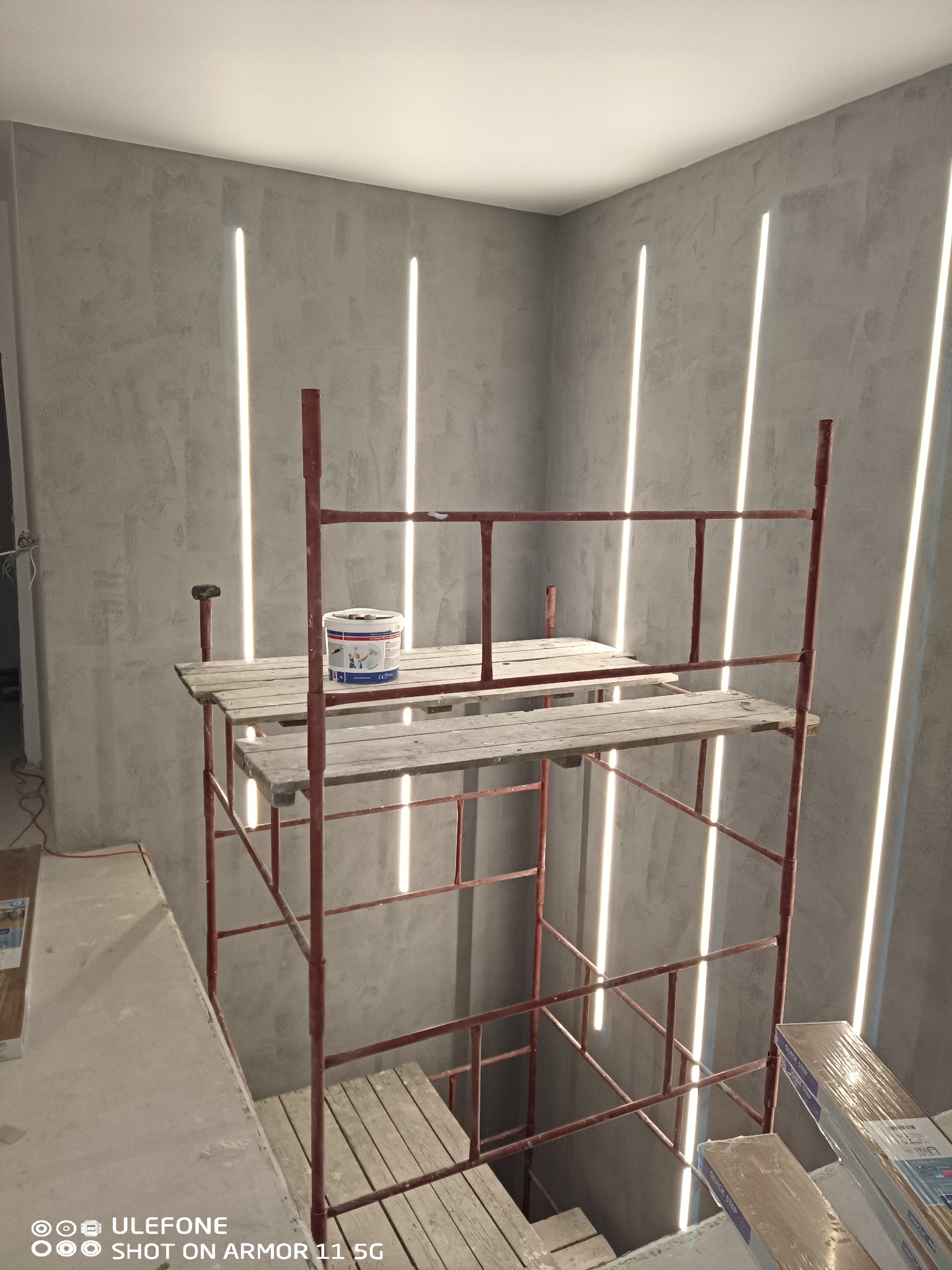 Szare ściany z dekoracyjnymi pionowymi pasami świetlnymi, czerwony metalowy ruszt, wiadro z farbą na drewnianej platformie rusztowania, częściowo widoczne panele podłogowe.