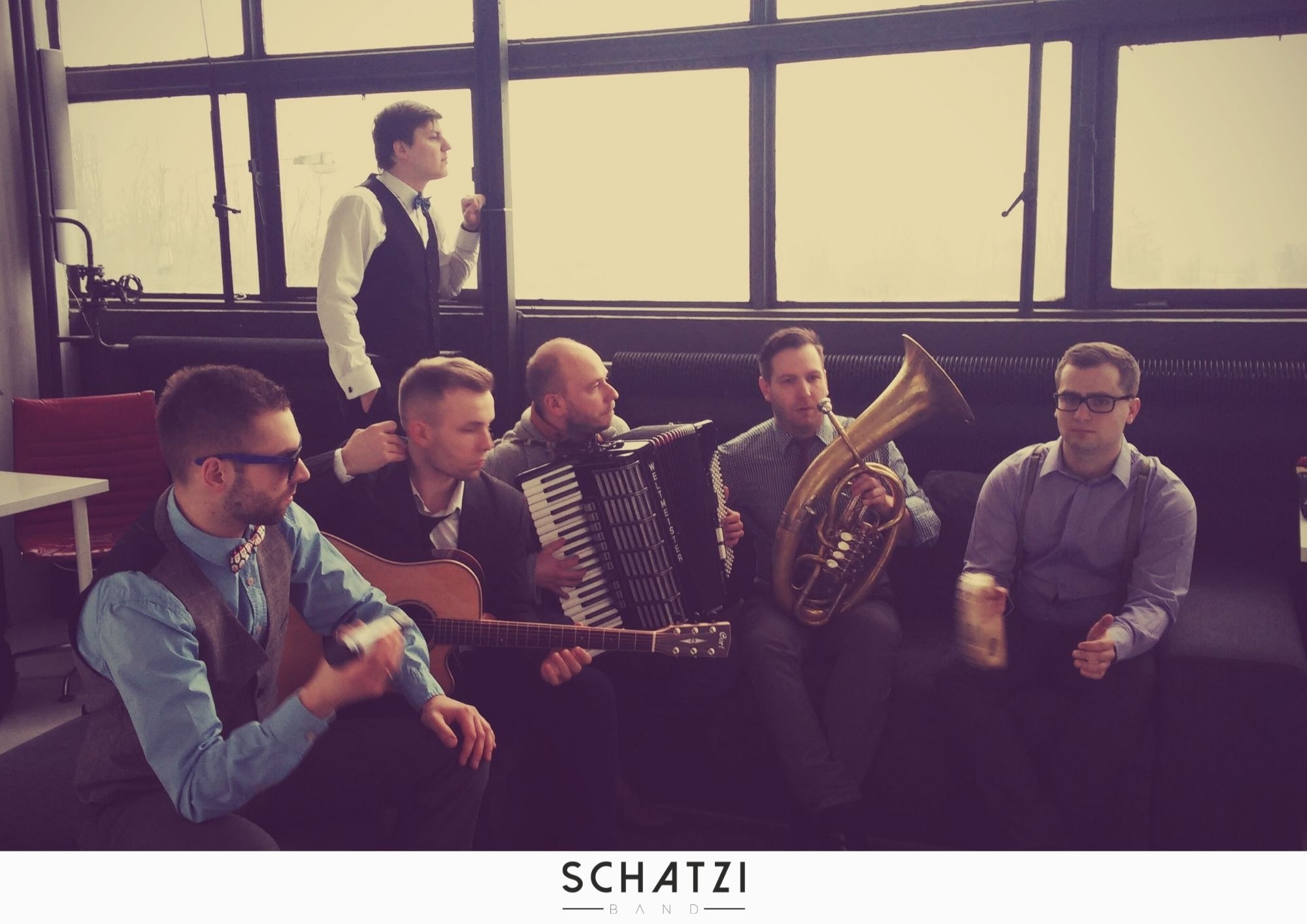 Sześcioosobowy zespół muzyczny w nieformalnym ustawieniu, z akordeonem, tubą, gitarą akustyczną; jeden członek w tle przy oknie, pozostali siedzą na kanapie.