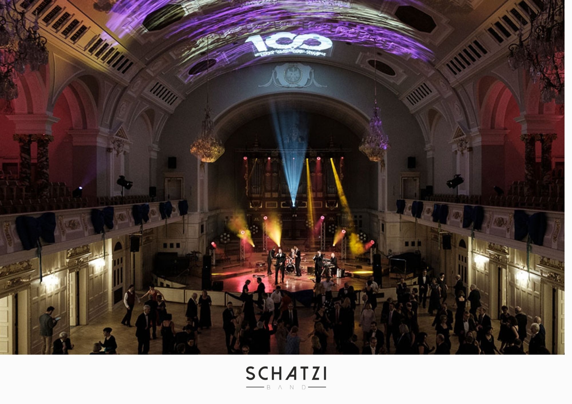 Zespół muzyczny na scenie w eleganckiej sali balowej z publicznością, oświetlenie sceniczne i logo zespołu wyświetlane na suficie.