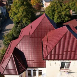 Widok z góry na dom z nowym, bordowym dachem z blachodachówki, z widocznymi obróbkami blacharskimi i rynnami, otoczony zielenią drzew i zabudową jednorodzinną.