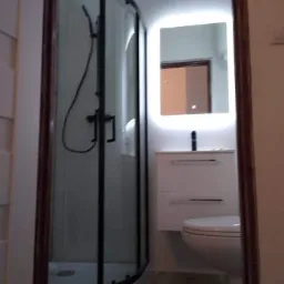 Wykończona łazienka z kabiną prysznicową z czarnymi profilami, podświetlanym lustrem nad umywalką i toaletą, widok zza futryny drzwi.