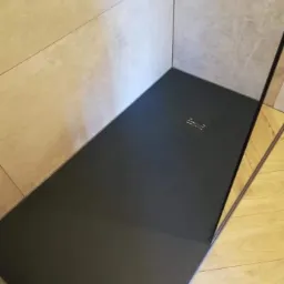 Nowoczesny brodzik bezprogowego prysznica z ciemnego kompozytu, otoczony jasnymi płytkami ceramicznymi z delikatnym wzorem marmuru, widoczny odpływ liniowy, ułożone panele podłogowe.