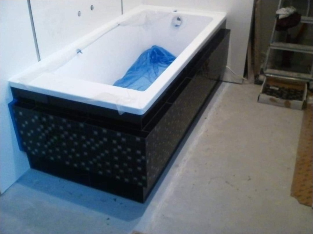 Wykończona obudowa wanny z czarnych płytek z delikatnym wzorem, wanna zabezpieczona folią, drabina i pudełko z elementami w tle, remont łazienki w trakcie.