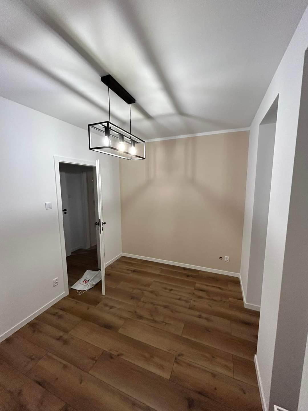 Wnętrze pokoju po remoncie: świeżo pomalowane ściany w odcieniach bieli i beżu, ułożona podłoga z paneli imitujących drewno, nowoczesna lampa wisząca z geometryczną oprawą, otwarte drzwi prowadzące...