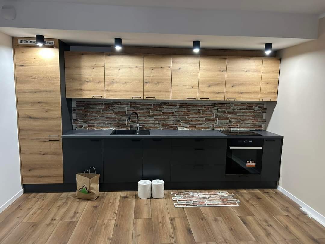 Nowoczesna kuchnia z drewnopodobnymi szafkami górnymi i czarnymi szafkami dolnymi, panel ścienny imitujący cegłę, oświetlenie punktowe, podłoga drewnopodobna, torba papierowa Leroy Merlin na podłodze.