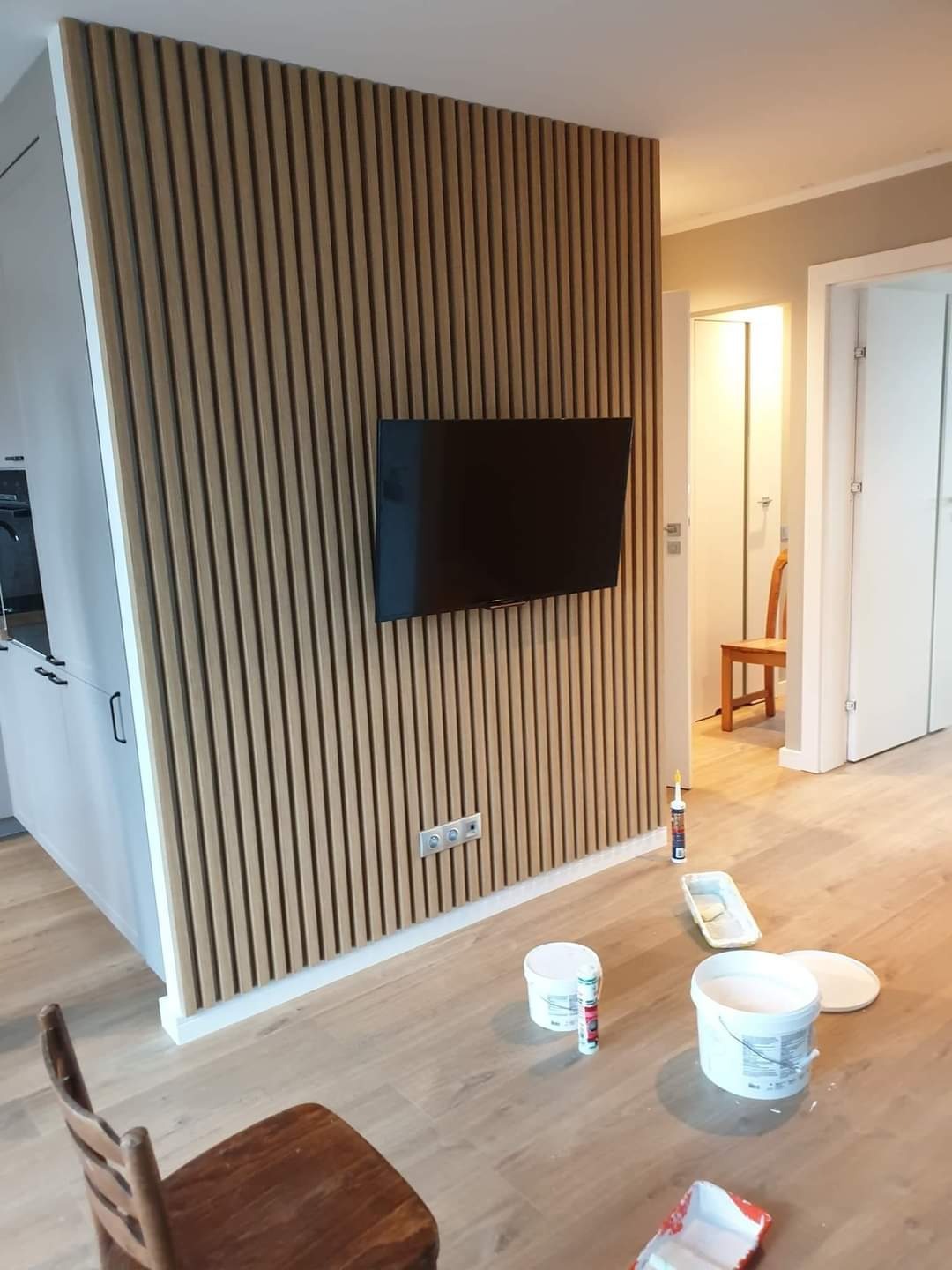 Ściana z drewnianych lameli z zamontowanym telewizorem w trakcie prac wykończeniowych, widoczne puszki z farbą i akcesoriami malarskimi na podłodze.