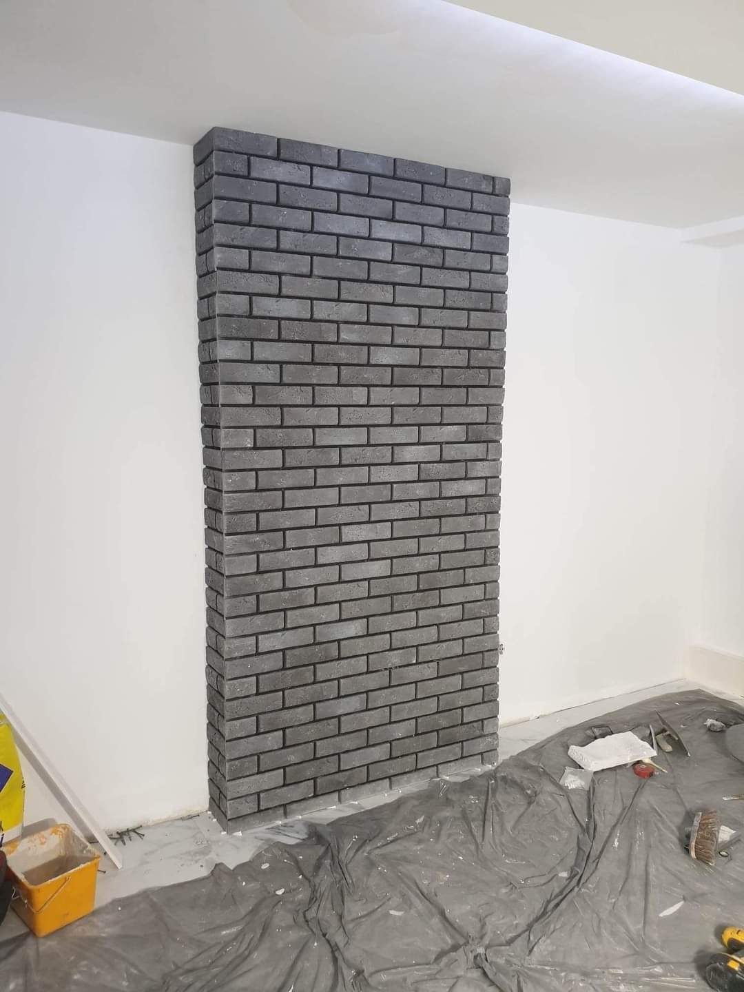 Ściana wykończona szarą cegłą klinkierową, widoczne narzędzia i folia ochronna na podłodze, etap prac wykończeniowych.
