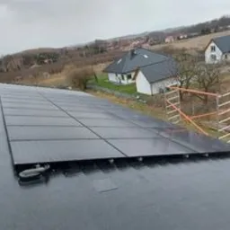 Panele słoneczne zamontowane na płaskim dachu budynku jednorodzinnego, widoczne rusztowanie, pochmurne niebo w tle.