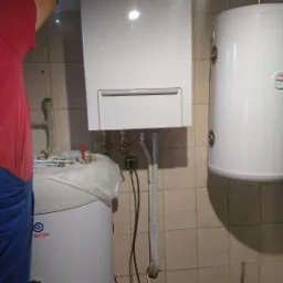 Instalacja wiszącego, białego, gazowego kotła grzewczego marki Samsung, widoczny także biały zasobnik ciepłej wody, instalator w czerwonej koszulce w trakcie montażu, beżowe płytki na ścianie.