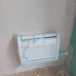 Biała skrzynka z instalacją hydrauliczną, częściowo zamaskowana folią ochronną, przymocowana do surowej ściany w trakcie montażu, widoczne niebieskie taśmy mocujące i fragment rury.